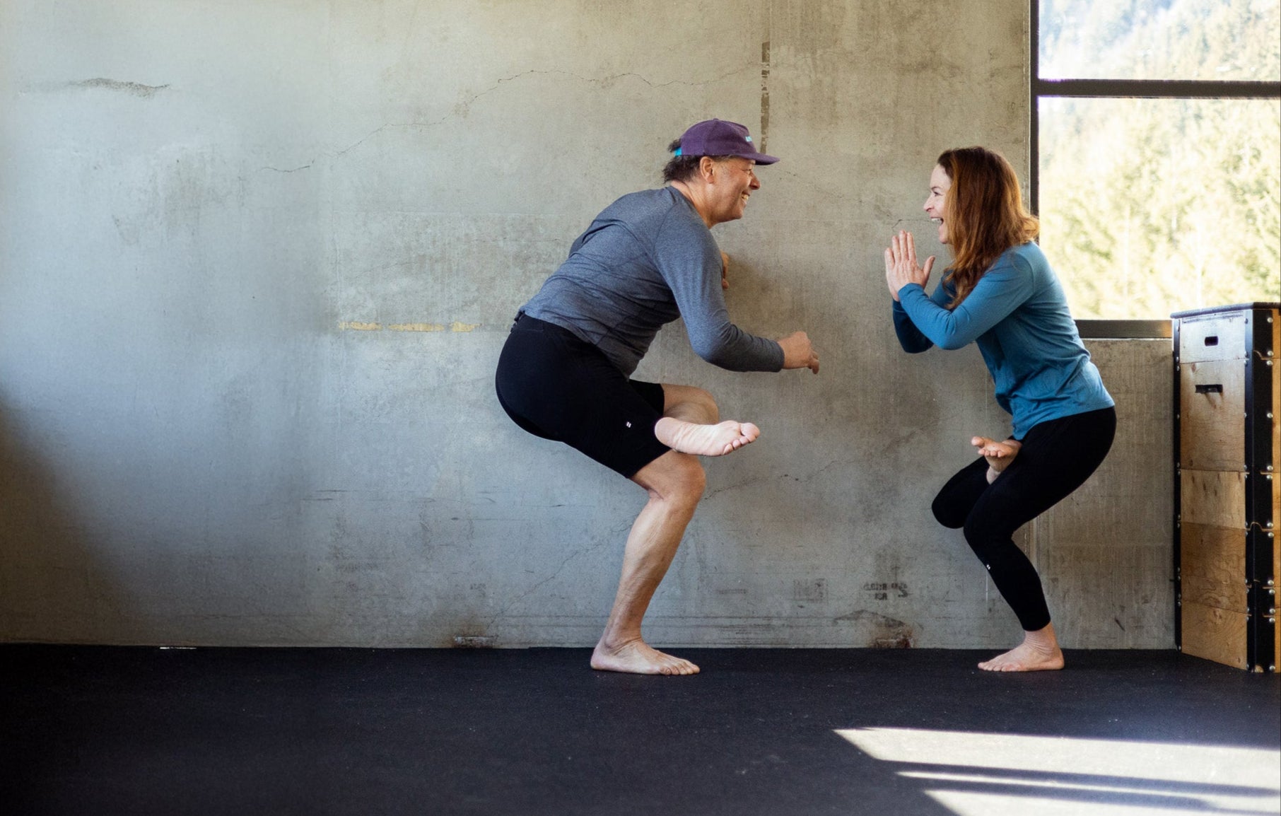 Two people performing a dynamic movement in a room with concrete walls and a window.